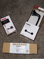 Three boxed bathroom hardware items including Delta Porter toilet handle (brown), Pfister Ladera matte black towel ring and rack, and a silver-tone towel ring.