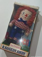 Raggedy Andy doll in original cardboard box, with visible plastic wrap covering the doll inside, box has colorful floral and sky designs, blue pants with buttons and checkered shirt on doll.