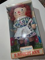 Original Raggedy Ann cloth doll with red yarn hair braided, wearing blue floral dress and white apron, inside original box.