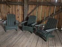 Three painted wooden Adirondack chairs in dark green, shown inside a barn on wooden floor, arranged in a semi-circle.