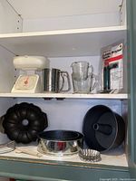 Photo showing white vintage style kitchen weigh scale, metal measuring cups, glass measuring pitchers, and boxed Crème Brûlée torch on a shelf.