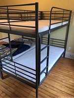 Metal twin bunk bed frame with two fitted white twin mattresses. View angled from side and slightly above, showing structure and bedding.