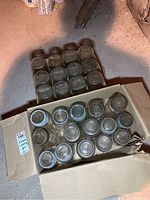 Two cardboard boxes containing a total of about 30 vintage glass canning jars in round and square shapes with some lids on top of the jars.