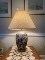 Full view of ceramic lamp on wooden table with lace doilies, lit, showing floral and bird design on blue base, pleated beige shade.