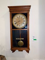 Full view of the regulator pendulum wall clock mounted on a wall showing wooden case, clock face and pendulum behind glass.