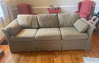 Front view of the beige Heritage Interiors couch showing three seat cushions, three back cushions, two arm pillows, and fabric skirted base on wooden floor.