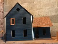 Front view of the wood siding doll house painted blue, showing window openings and a brown shingle roof section on the right side.