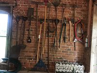 Photo of garden tools (shovels, rakes, pruners, axe, saws) hanging on brick wall with metal coated hanging baskets below and green garden hose coiled up on floor.