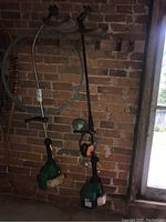 Two green gas-powered Weed Eater trimmer units hanging on a brick wall with long shafts and loop handles. A reel of orange replacement trimmer cord is visible on one unit. Ear protectors and a roll of wire also included.
