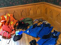 Photo showing five orange life jackets, pair of black water shoes, several blue and black wetsuits, snorkeling gear including goggles and flippers spread on wooden floor.
