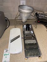 Photo showing a white and stainless steel mandolin slicer with black hand guard next to a conical stainless steel strainer held by a tripod wire stand