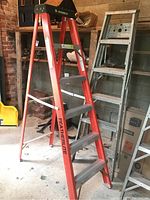 Two step ladders displayed upright, one orange Featherlite fiberglass ladder and one aluminum ladder against the wall in a garage.