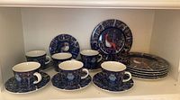 Wide view showing 6 teacups paired with 6 matching saucers and 6 dessert plates stacked behind, all with colorful bird and floral designs on navy background.