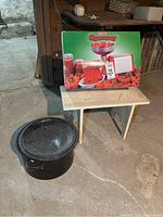 Original box of Imperia Spremy tomato squeezer displayed on a small wooden table along with the large black roasting pot on the floor.