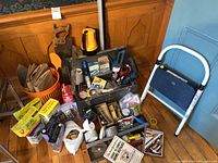 Overview of the lot showing metal stepping stool, wooden tool chest, saw, corded drill, various hand tools, rat poison chemicals in yellow boxes, flashlights, and assorted hardware on floor.