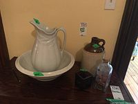Wide shot of full lot showing wash basin and pitcher, crock, decorative glass bottle with floral design, and small candleholder on wood surface against yellow wall.