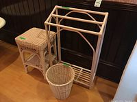 Photo of all three items: wicker side table, wicker waste basket, and rattan towel/blanket rack grouped together on a wooden floor against a dark wood paneled wall.