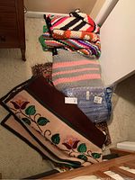 Photo showing four crocheted afghans with various colorful striped patterns, one folded woven floral rug/blanket, and a packaged blue and purple blanket.