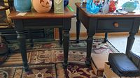Two matching wooden end tables with reddish-brown tops and dark green turned legs. Positioned next to each other on patterned rugs with visible scratches.