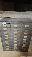 Front view of the 27-drawer metal file box cabinet showing labeled drawers and dust on the surface.