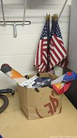 Wide shot showing multiple large American flags on poles leaning against wall, smaller flags inside glass jar, and additional flags inside paper bag on table.