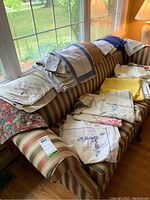 Multiple vintage linens including napkins, placemats and tablecloths folded and laid across the back of a striped couch by a window.