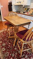 Full view of wooden rectangular table with two matching chairs with red seat cushions in a kitchen setting on a decorative rug.