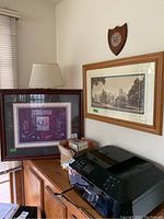 Photo showing golf print framed in dark wood and vintage university photo framed in light wood above a desk with a lamp and printer
