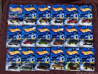 Top view of 18 sealed Hot Wheels cars in blue carded packages arranged in rows on a dark surface, showing front packaging and some model names.