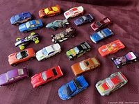 Top-down angled photo showing all 20 vintage Hot Wheels and die-cast vehicles arranged closely on a purple fabric surface.