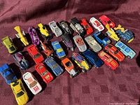 Overview photo showing 31 vintage die-cast vehicles arranged on a maroon cloth surface, highlighting various colors, styles and models including race cars and utility vehicles.