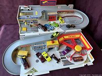Full view of the vintage Hot Wheels service station playset with 14 die-cast vehicles arranged on and around it, showing ramps, car wash area, and storefront details.