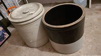 Wide view of two ceramic crocks placed side by side on a concrete floor. One crock has a lid with a crack.