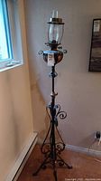 Full view of tall antique gas floor lamp with wrought iron scroll base, hammered copper tank, and glass chimney.