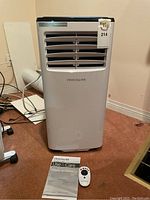 Full front view of Frigidaire portable air conditioner with remote and manual placed on the floor in front.