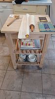 Full view of wood kitchen island with butcher block top with wooden mallet, large rolling pin, vintage cookbooks, kitchen towels, and ceramic bowls on shelves below.