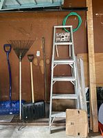 Garage tools including aluminum ladder, shovels, rakes, hose, and wood leaning against a brown wall with visible stains and wear.