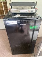 Black Hamilton Beach mini refrigerator with stickers on the door, some surface wear visible.