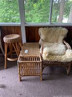 Photo of rattan armchair covered with white sheepskin, rectangular coffee table with woven top and built-in magazine holder, and round rattan bar stool, all placed together on carpeted floor near window with wood paneling background.
