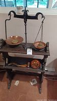 Full view of vintage cast iron balance scale displayed on a wooden two-tier shelf with copper pans and brass weights.