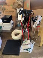 View of red Hugo walker folded with black seat and wheels, black cushion and white toilet seat riser nearby on the floor.