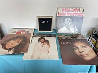 Photo showing five vinyl record albums laid flat on a blue surface, featuring Barbra Streisand and Louis Armstrong albums with visible cover artwork.