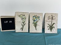 Three ceramic floral plaques showing Orchid, Iris, and Amaryllis flowers. Each plaque is rectangular and textured with painted raised floral details.