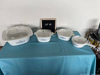 Set of four blue cornflower Corningware casserole dishes with lids on blue cloth-covered table.