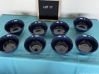 Eight dark blue porcelain cereal bowls arranged in two rows on a light blue cloth with the black and white LOT 17 sign behind them.