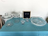 Photo showing two clear glass canisters with lids, two small taper candle holders, and a round serving plate arranged on a blue cloth-covered surface.