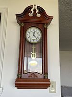 Front view of wooden great grandmother clock mounted on wall with visible pendulum and clock face showing time.