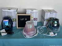 Three Scentsy warmers (two blue, one pink) shown in front of their original white packaging with decorative bands, electrical cords included.