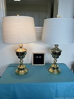 Pair of brass-colored table lamps with white drum shades and unique finials, one horse-shaped and one butterfly-shaped.
