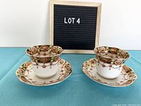 Two cups with matching saucers arranged side by side with a letter board in background showing 'LOT 4'. Cups and saucers display ornate green and brown patterns with gold trim.